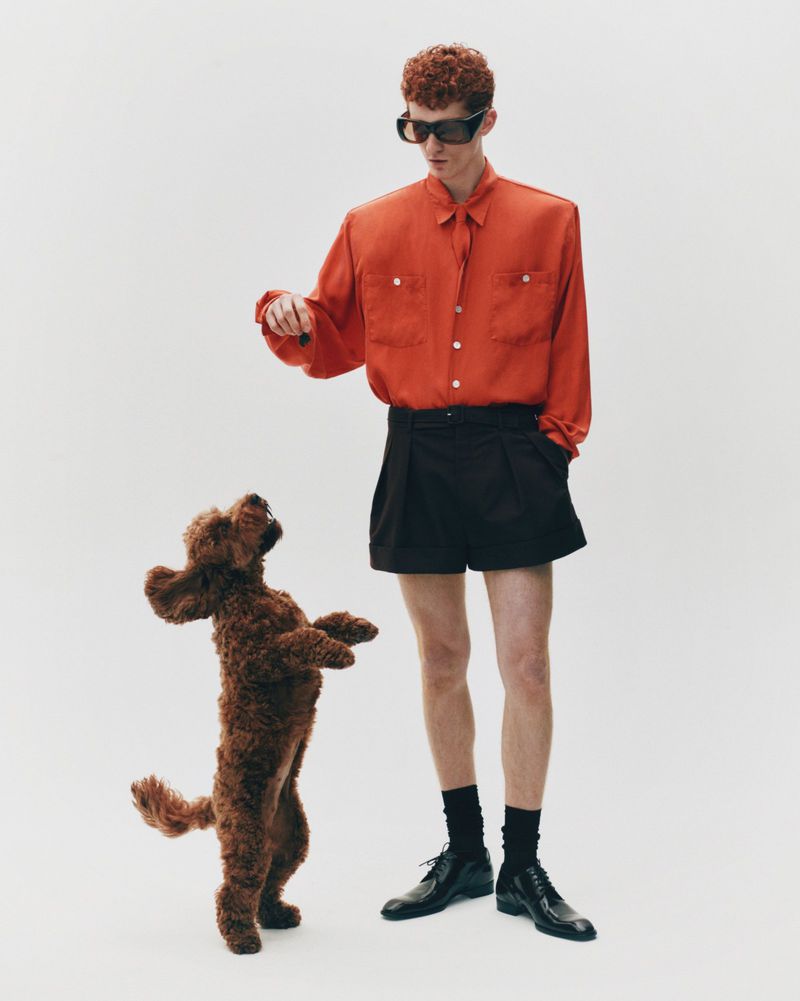 Stylish man in orange shirt and black shorts poses with a brown poodle on a white background in a fashion editorial shot.