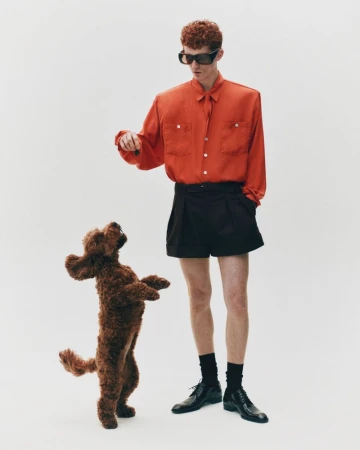 Stylish man in orange shirt and black shorts poses with a brown poodle on a white background in a fashion editorial shot.