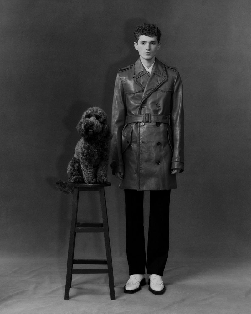 Monochrome portrait of a curly-haired young adult male in a leather trench coat standing next to a poodle on a wooden stool.