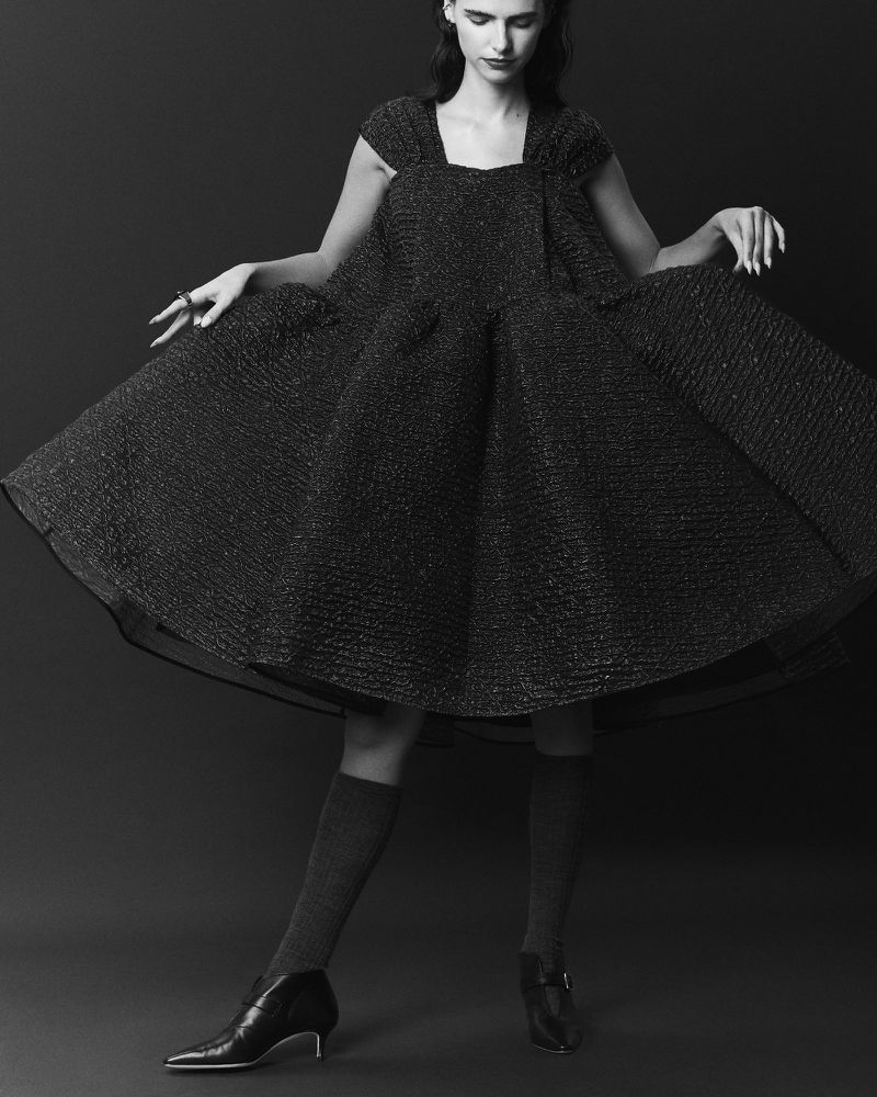 Monochrome fashion shot of a slender woman in a dark textured dress with knee socks and low heels against a dark grey background.