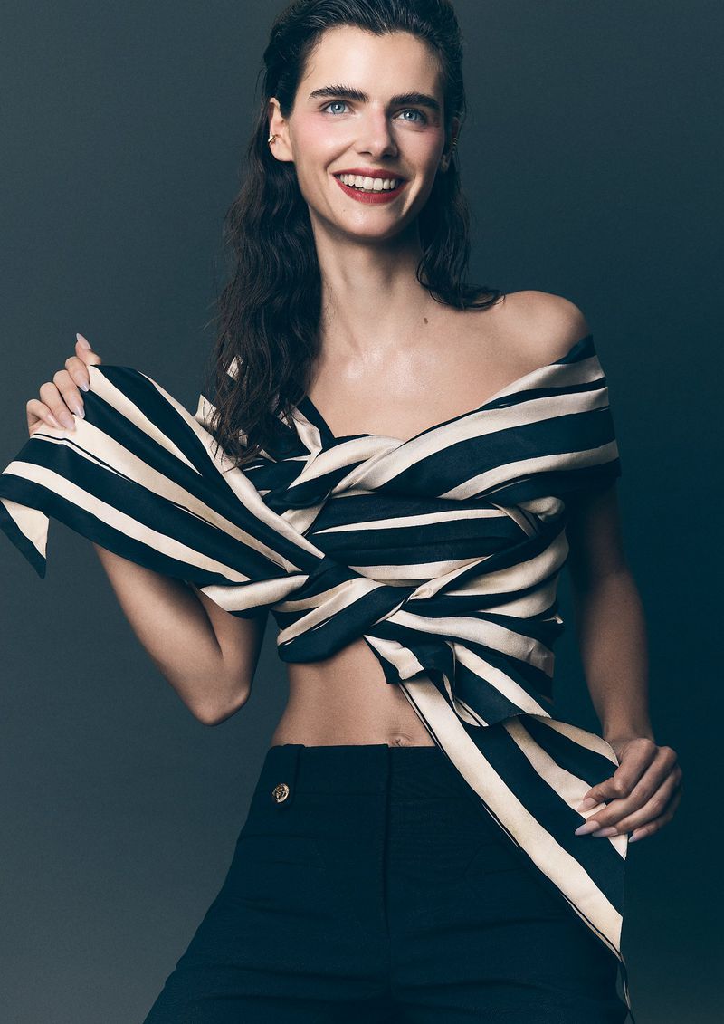 Portrait of a smiling woman in a striped top and black pants against a grey background, showcasing a fashion-forward editorial style.