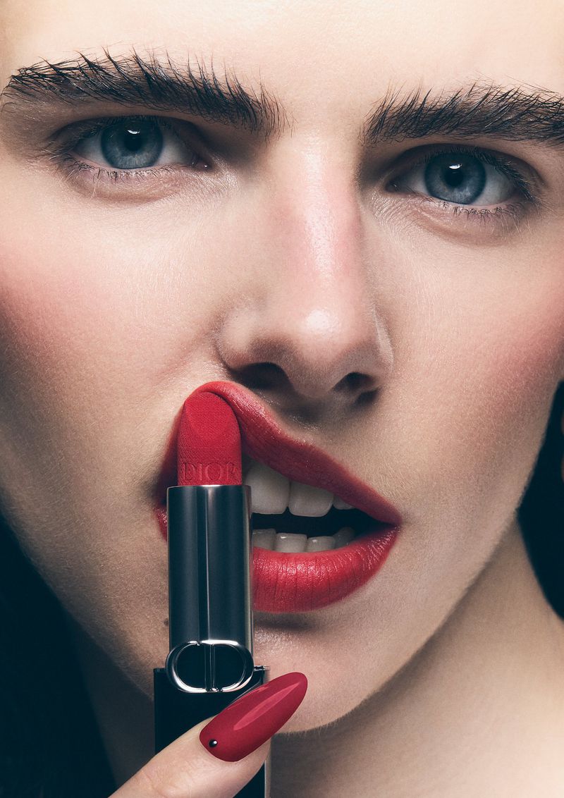 Close-up portrait of a fair-skinned woman holding a red lipstick tube, with matching red lips and nails against a dark backdrop.