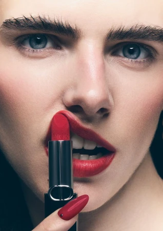 Close-up portrait of a fair-skinned woman holding a red lipstick tube, with matching red lips and nails against a dark backdrop.