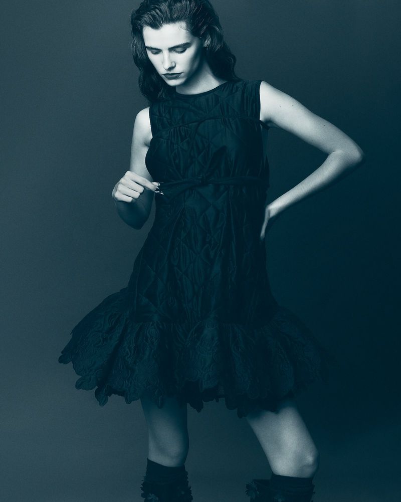 A young adult woman poses indoors, wearing a short black dress and dark boots, with a grey background in a fashion studio shoot.