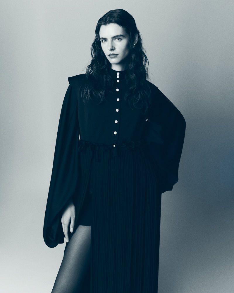 A full-length portrait showcases a poised woman in a sophisticated black dress with a thigh-high slit, set against a cool grey backdrop.
