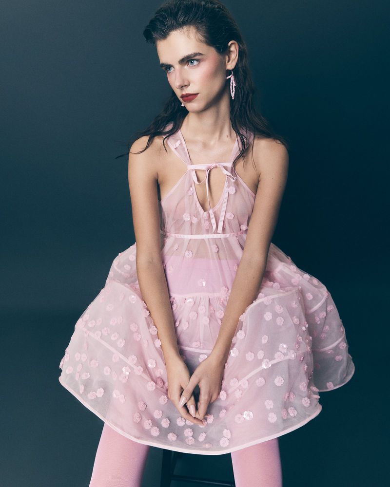 A woman in a pink dress poses in a studio, showcasing a light and airy fashion style with delicate floral details and pink tights.