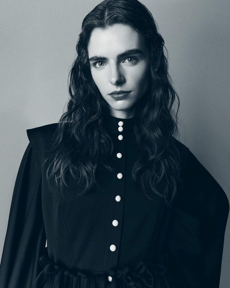 Monochromatic portrait of a woman with long, wavy hair, wearing a high-necked dress with pearl buttons in a studio setting.
