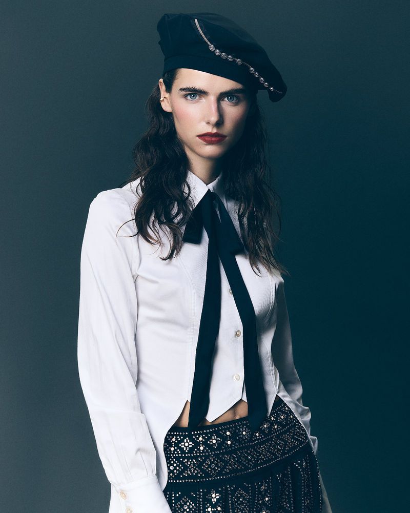 A woman poses indoors wearing a white collared shirt, black beret, and decorated skirt, captured in a medium shot with a solid backdrop.