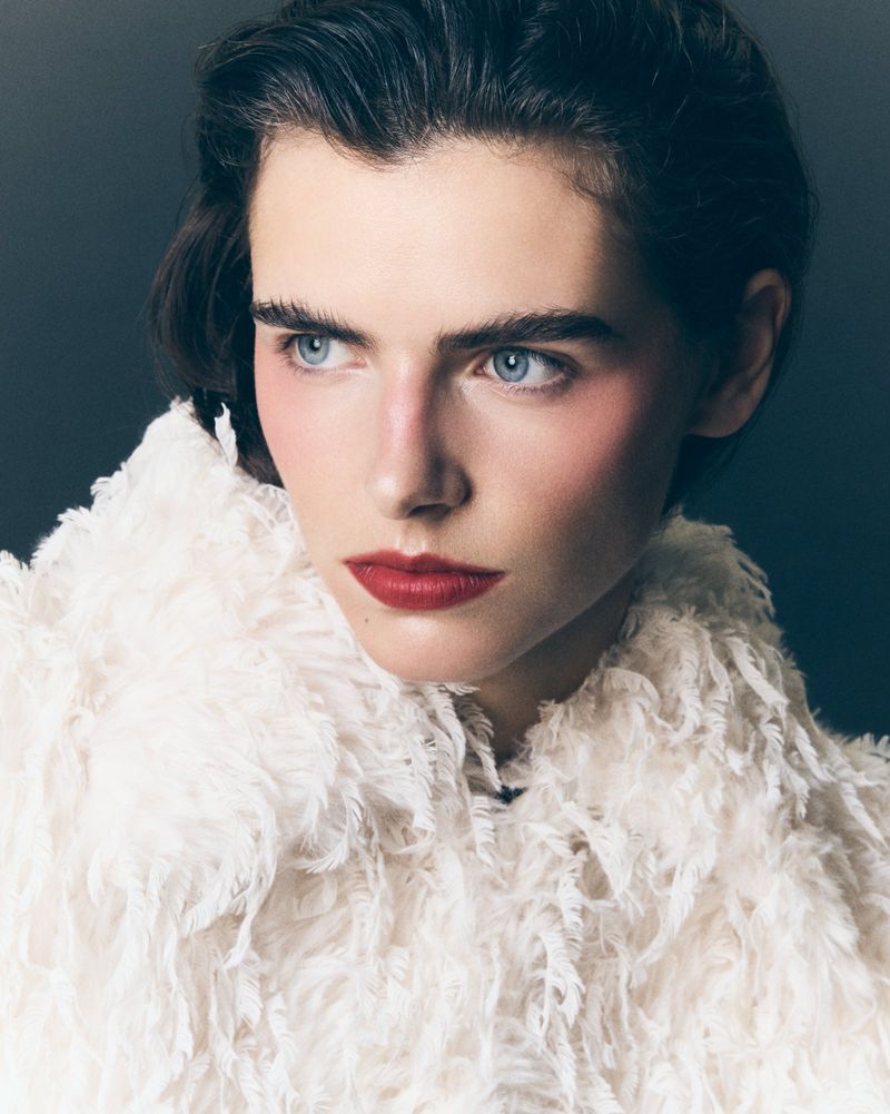 Close-up portrait of a fair-skinned adult woman with blue eyes and dark hair, wearing a white feathered garment against a dark background.