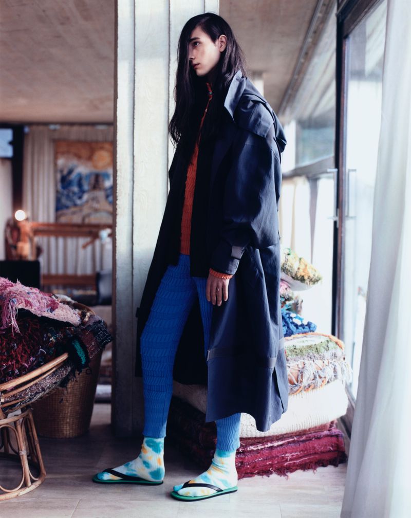 Stylish individual with long dark hair in a black coat, blue leggings, and colorful socks, standing indoors near a window with filtered light.