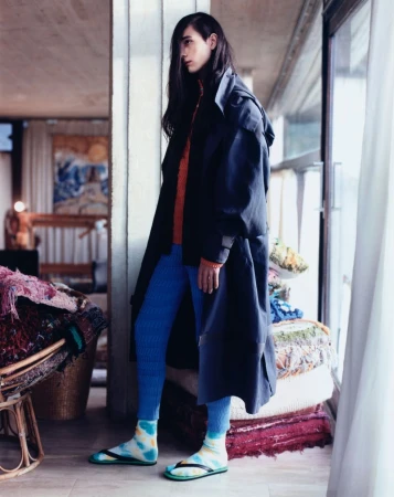 Stylish individual with long dark hair in a black coat, blue leggings, and colorful socks, standing indoors near a window with filtered light.