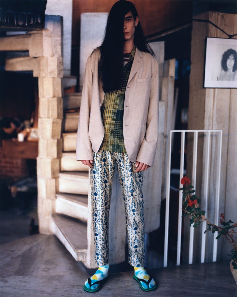Standing adult with long dark hair wearing a blazer, patterned shirt and pants, and sandals inside a room with a spiral staircase.