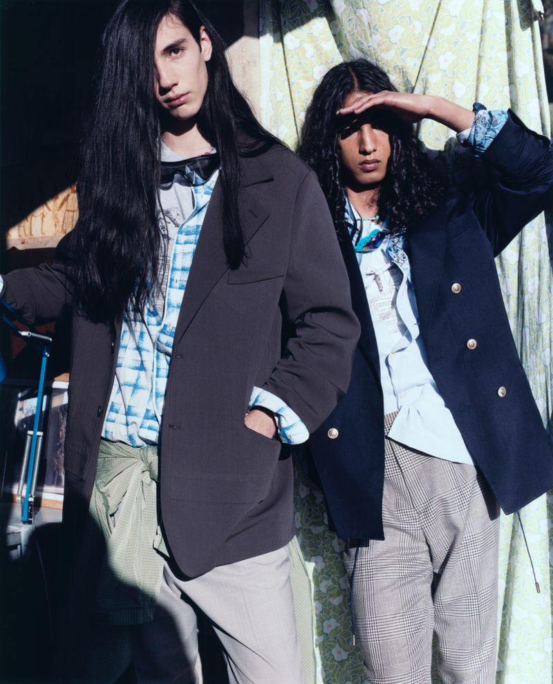 Two young men pose in blazers and button-down shirts, one shielding his eyes, against a backdrop of green patterned fabric.