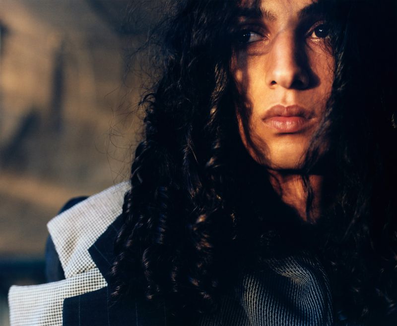 Portrait of a person with dark curly hair, looking to the side and wearing a blazer with light and dark shadows.