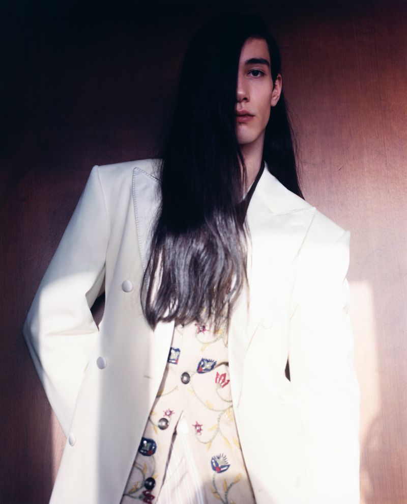 Portrait of a stylish adult male with long dark hair, wearing a white double-breasted blazer with a patterned shirt, against a blurred background.