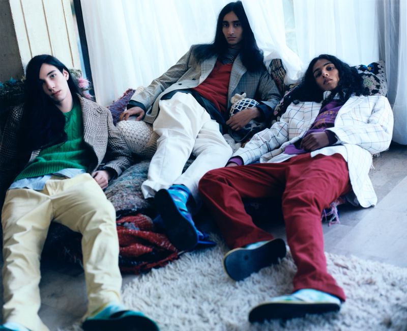 Three dark-haired adult male models in blazers and casual pants pose indoors on a plush rug with throw pillows in a fashion editorial shot.