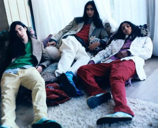 Three dark-haired adult male models in blazers and casual pants pose indoors on a plush rug with throw pillows in a fashion editorial shot.