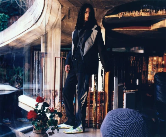 Fashionable adult male model with long hair poses in a modern home with large windows, wearing a dark blazer and trousers with colorful shoes.