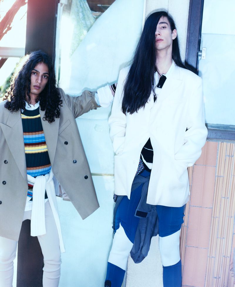 Two people in stylish casual wear pose outdoors in daylight. One wears a tan blazer and the other wears a white blazer and colorful pants.