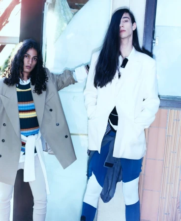 Two people in stylish casual wear pose outdoors in daylight. One wears a tan blazer and the other wears a white blazer and colorful pants.