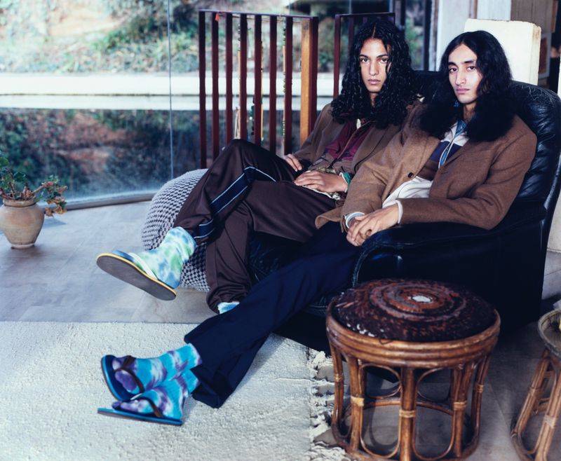 Two adult males with long hair seated indoors, wearing blazers, trousers, and socks with sandals, gazing at the camera in a relaxed setting.