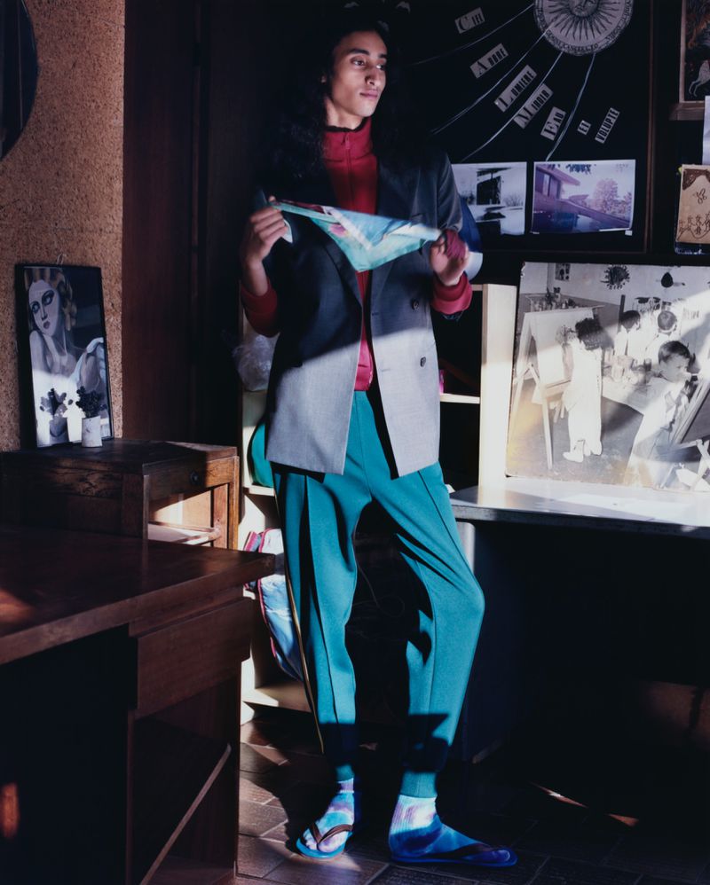 Fashion shot of a young adult in a grey jacket and teal pants standing indoors with artwork on the wall in the background.