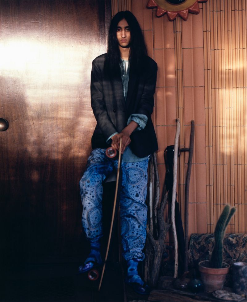 A young adult with long dark hair sits in a studio wearing paisley pants, a blazer, and roller skates, leaning on a cane with a wooden door.