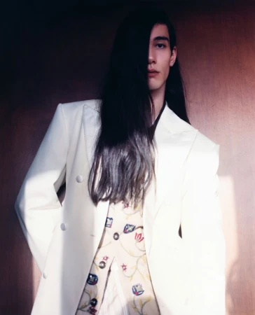 Model with long dark hair wearing a white double-breasted blazer and patterned vest against a wooden wall
