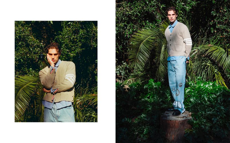 Male fashion model wearing a beige cardigan, light blue shirt, and jeans with peace symbols in an outdoor nature setting.
