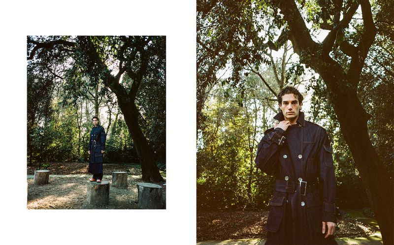 A man in a dark trench coat poses outdoors surrounded by trees, showcasing fashion and nature photography, captured in natural light.