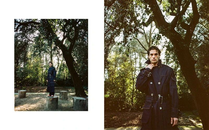 A man in a dark trench coat poses outdoors surrounded by trees, showcasing fashion and nature photography, captured in natural light.