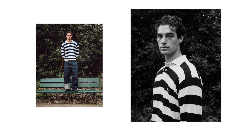 A young man stands on a bench in a striped shirt and jeans in an outdoor fashion editorial, captured in a bright, natural light.