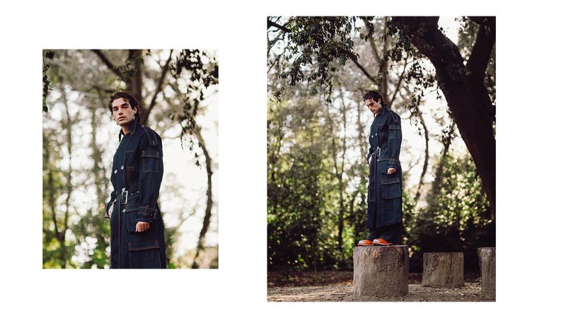 A man in a long blue coat poses in a forest with dappled sunlight, standing on a tree stump for an outdoor fashion shot.