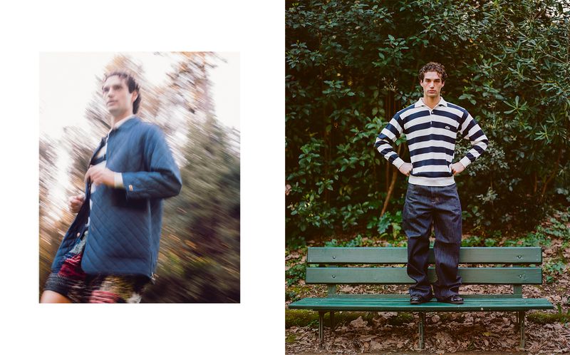 A male fashion model poses on a park bench in a striped sweater, while another blurry male figure walks amidst foliage in a blue coat.
