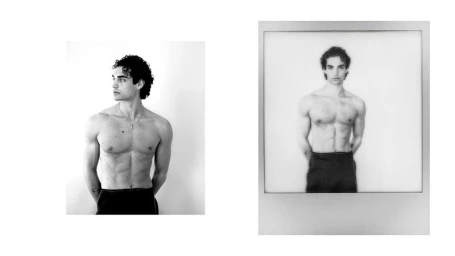 Black and white portrait of a shirtless young man with dark curly hair and a pendant necklace against a plain white wall