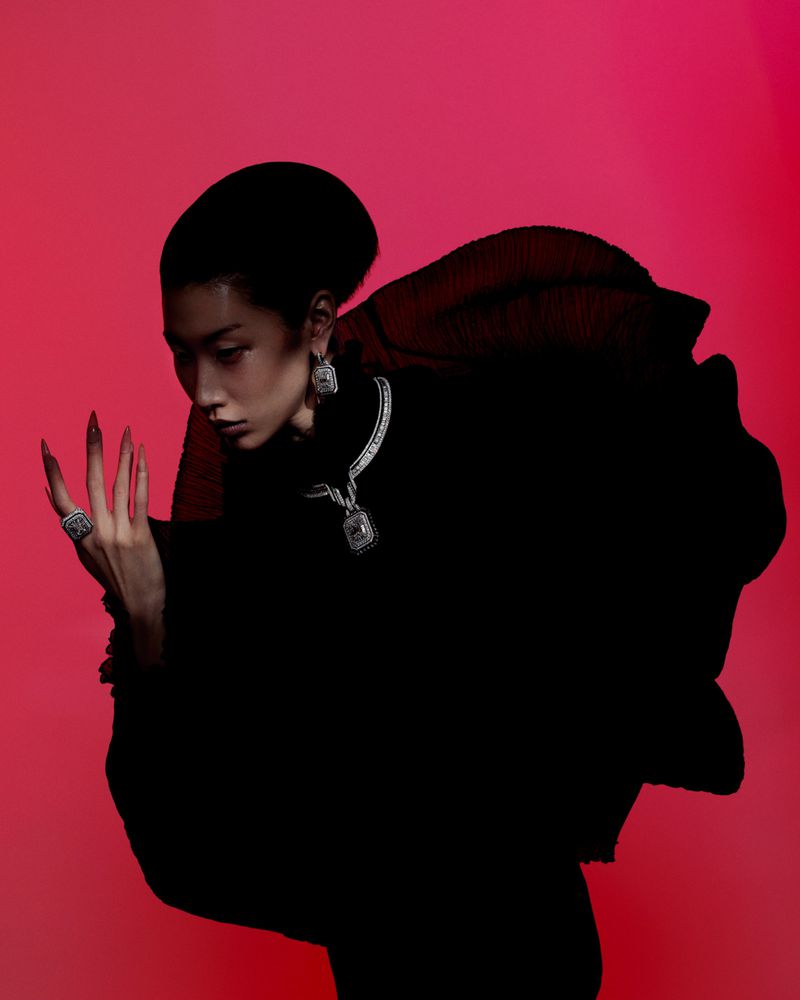 A woman with long nails wears a black dress and silver jewellery against a red background in a beauty and fashion setting.