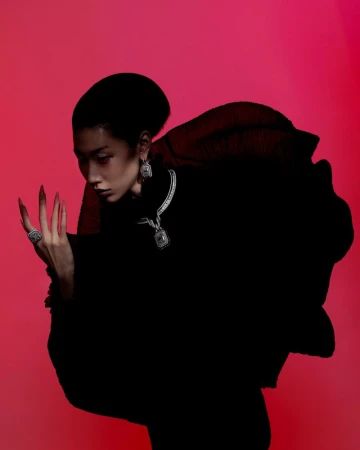 A woman with long nails wears a black dress and silver jewellery against a red background in a beauty and fashion setting.
