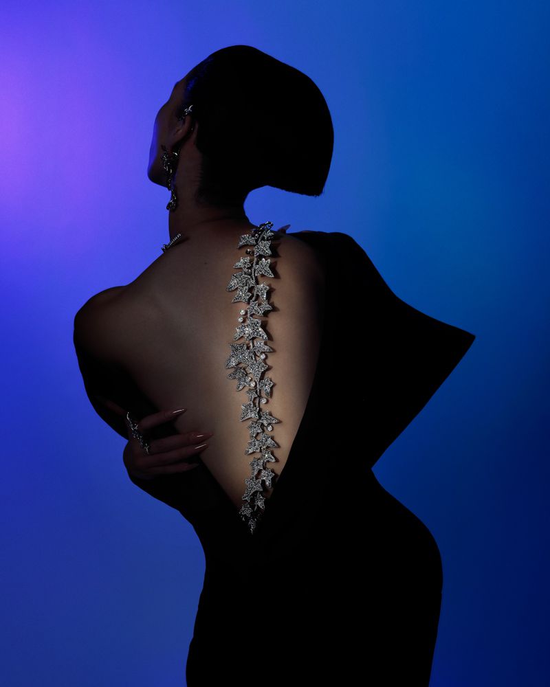 Adult woman wearing a black dress with a floral gemstone spine piece, jewelry, and updo hairstyle set against a gradient blue and violet background.