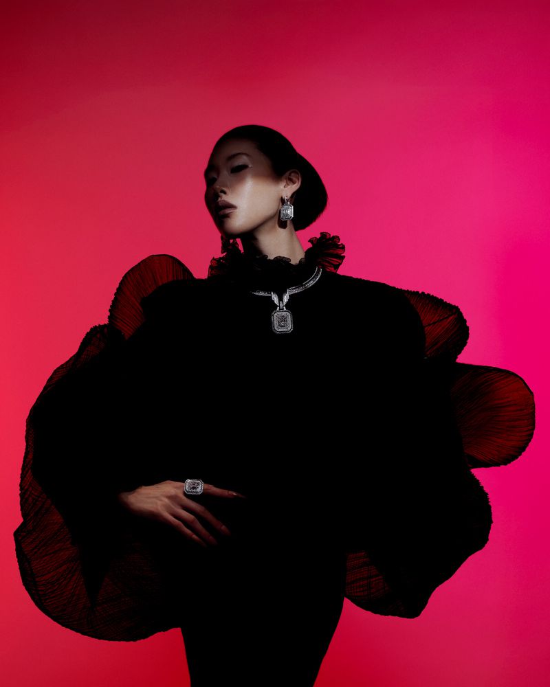 Elegant woman wearing large black ruffled top with statement jewelry, in front of a vibrant, gradient pink background in studio lighting.
