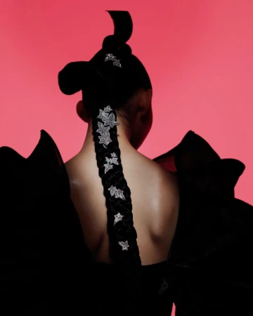 Model with black braided hair decorated with silver stars posing against a pink background wearing a ruffled black dress.