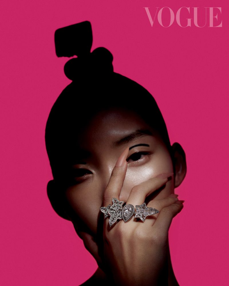Close-up fashion shot of an Asian woman with dramatic eye makeup and rings with star and teardrop shapes against a hot pink backdrop.