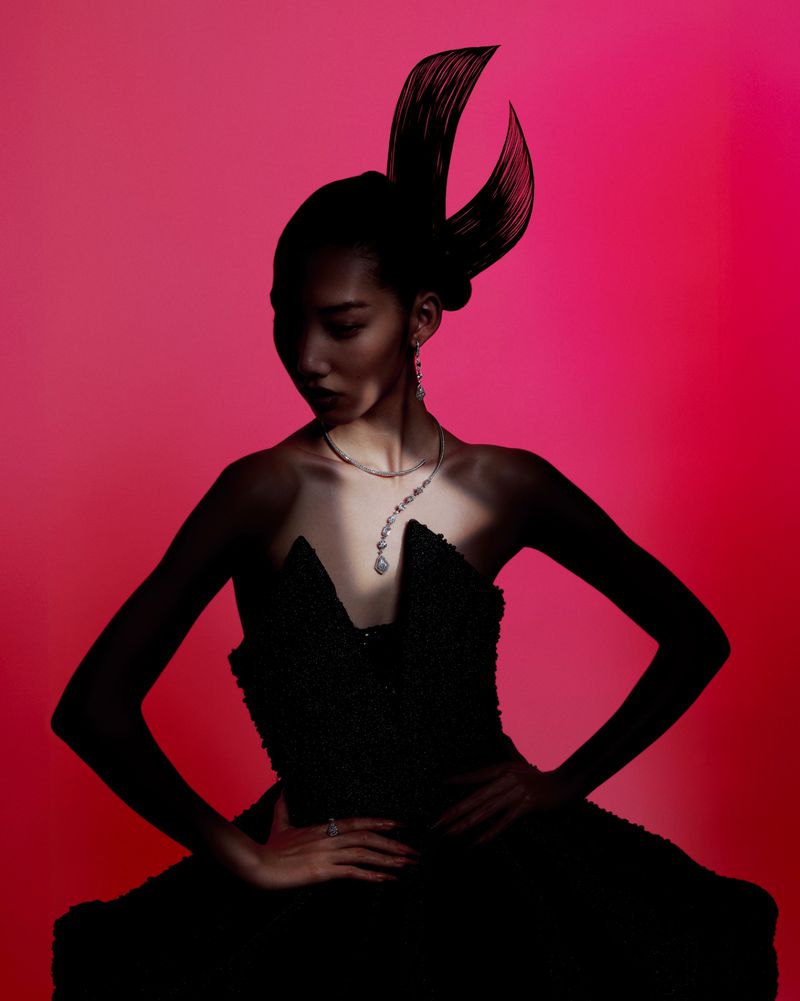 Woman in strapless gown and dramatic headpiece poses against a pink background. She wears a statement necklace and has her hands on her hips.