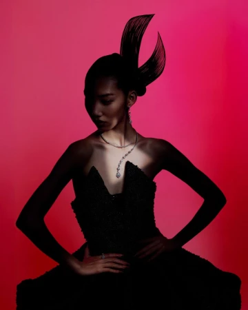 Woman in strapless gown and dramatic headpiece poses against a pink background. She wears a statement necklace and has her hands on her hips.