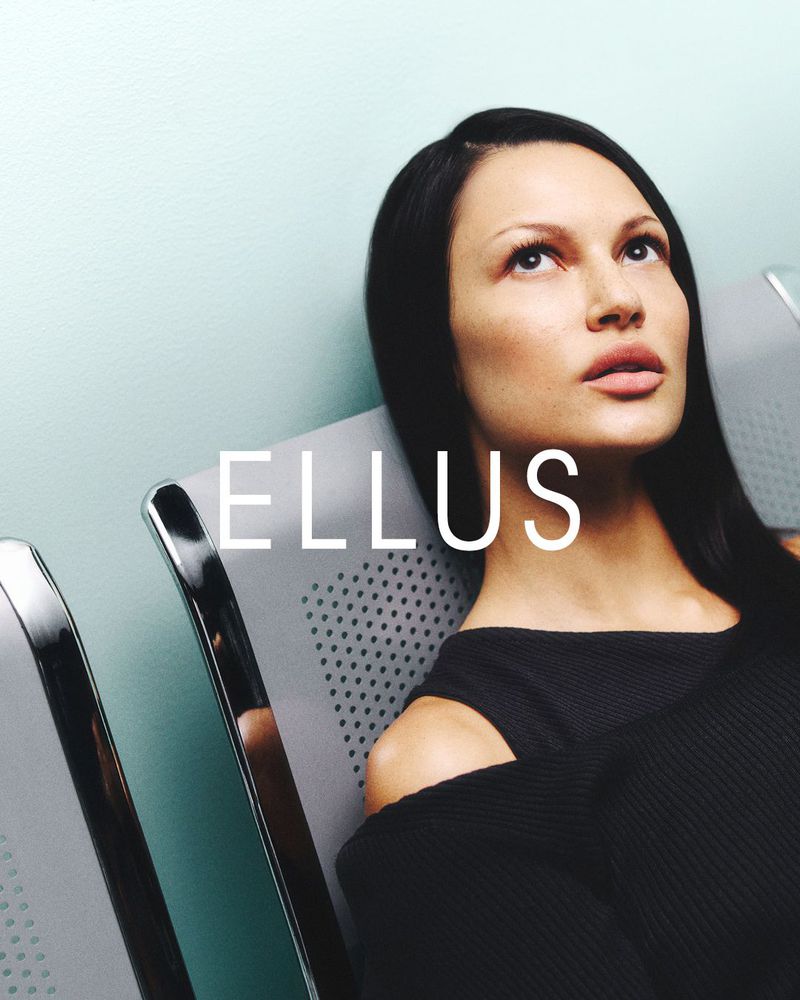 Portrait of a contemplative adult woman seated in a waiting area, wearing a black sweater, looking upward near a wall and gray chairs.