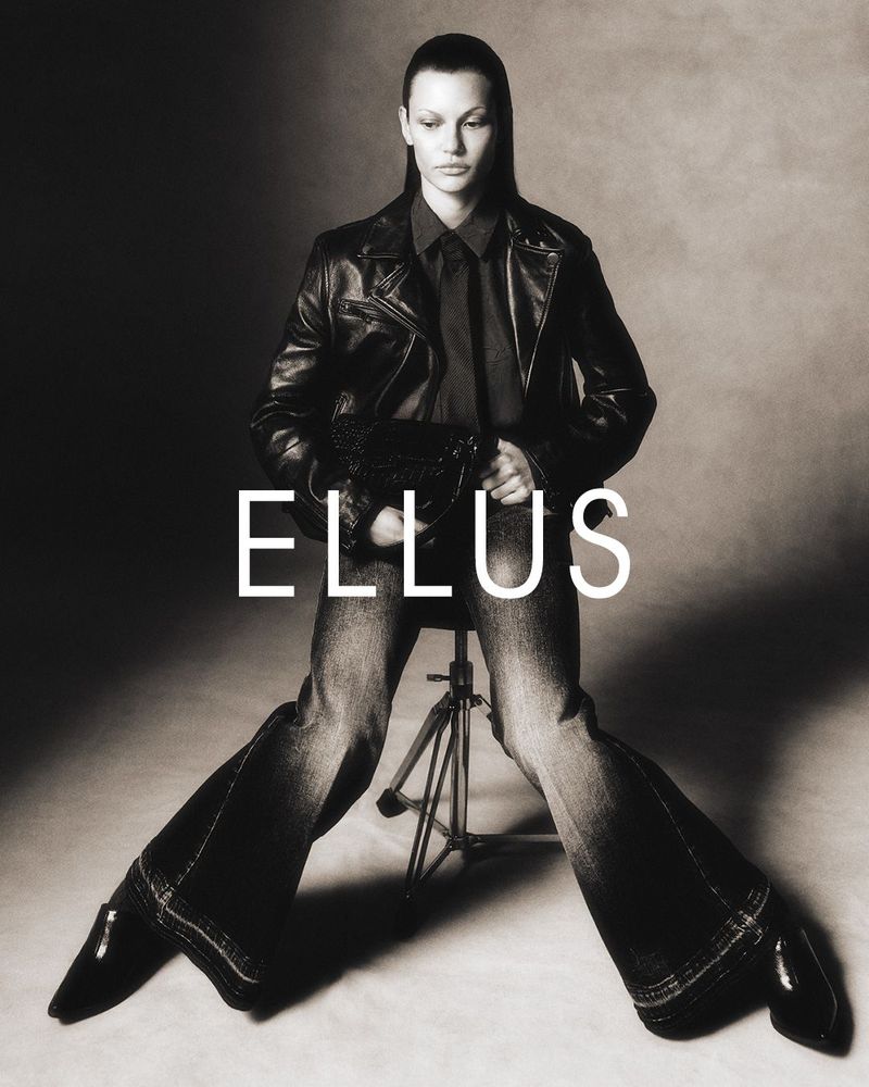 Monochrome fashion shot of a model in a black leather jacket, jeans, and loafers, seated on a camera tripod, set against a plain backdrop.