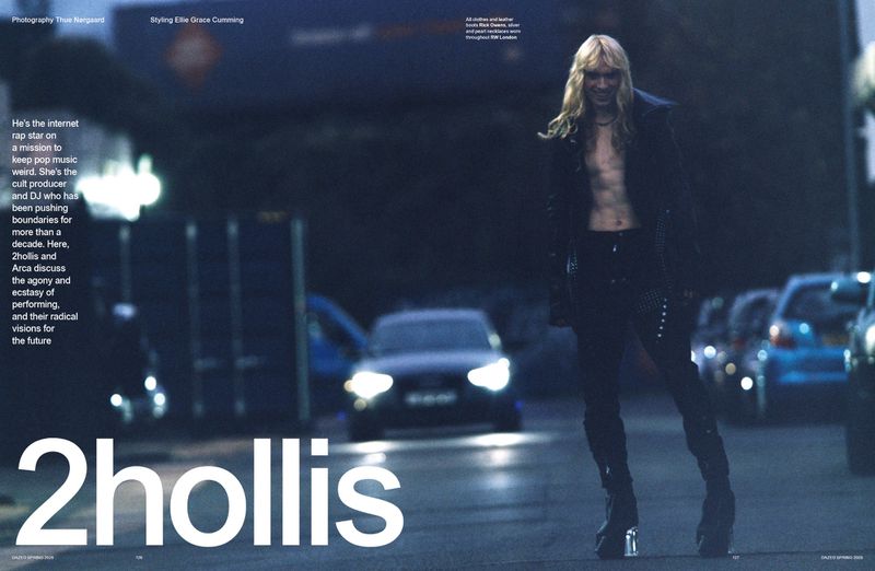 A person stands on a dark street at night, wearing black leather, with cars parked and buildings in the background, magazine editorial.