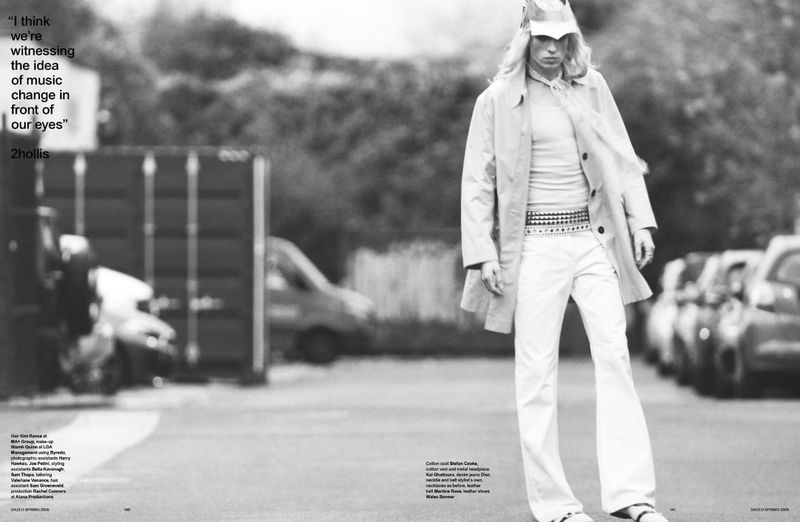 Fashion shot of a blonde male model in a beige trench coat and white jeans walking down a street with parked cars and buildings.