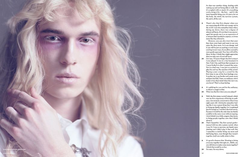 Portrait of a fair-skinned male with platinum blonde curly hair, wearing makeup and a collared shirt in a studio setting.