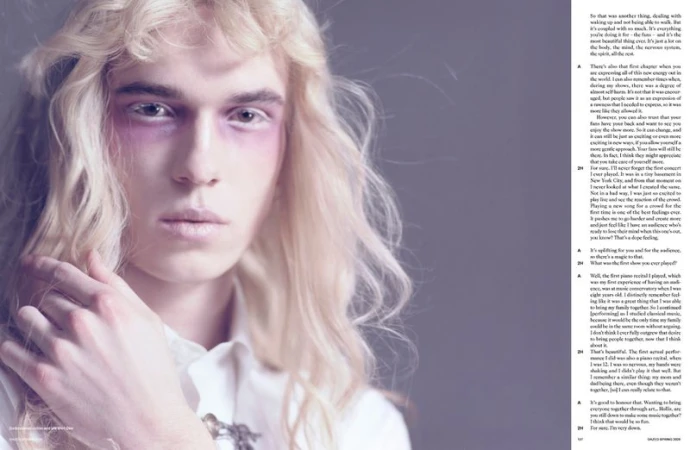 Portrait of a fair-skinned male with platinum blonde curly hair, wearing makeup and a collared shirt in a studio setting.