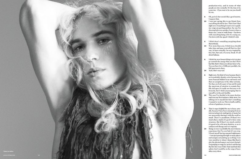 Monochrome portrait of a young adult with blonde hair. Woman is posing with arms raised and a textured black garment around shoulders.